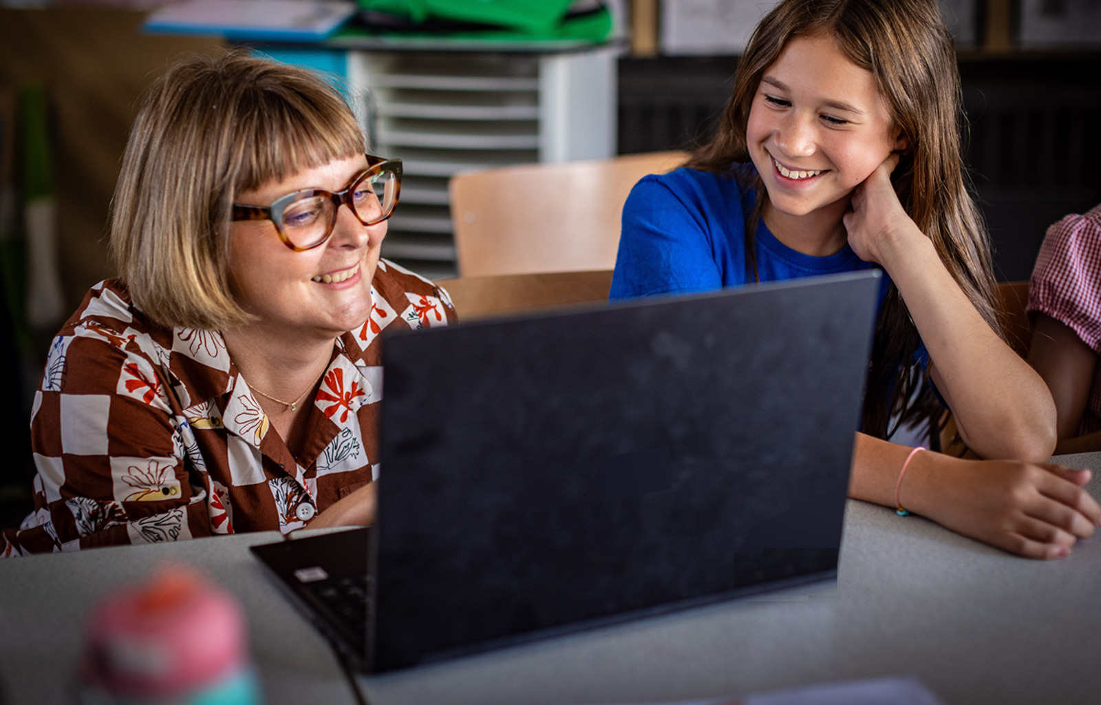 A student and a teacher sit together, looking at a laptop and smiling.