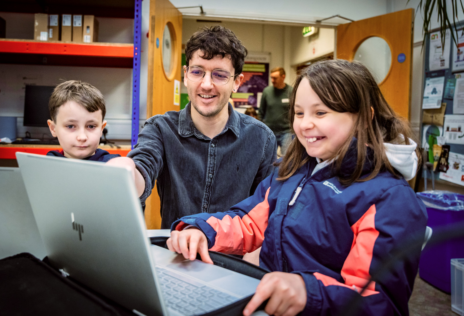 A teacher sits between two students. They are all looking at a laptop screen and smiling.
